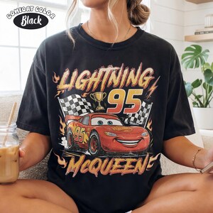 Puede incluir: Una camiseta negra de manga corta con un gr&aacute;fico de un coche de carreras rojo con el n&uacute;mero 95, un trofeo dorado y banderas a cuadros. El texto dice LIGHTNING 95 MCQUEEN en una fuente amarilla y naranja desgastada con llamas.