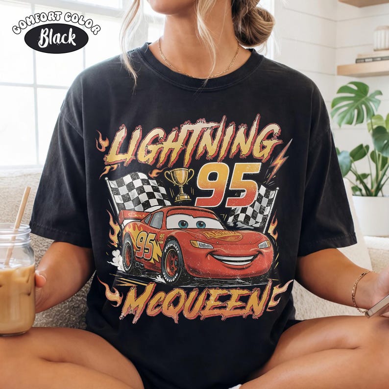 Puede incluir: Una camiseta negra de manga corta con un gr&aacute;fico de un coche de carreras rojo con el n&uacute;mero 95, un trofeo dorado y banderas a cuadros. El texto dice LIGHTNING 95 MCQUEEN en una fuente amarilla y naranja desgastada con llamas.