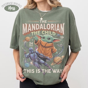 May include: A moss green t-shirt featuring a graphic print of The Mandalorian and The Child in space. The design includes planets, stars, and the text THE MANDALORIAN, THE CHILD, 24 JULY, 12c, and THIS IS THE WAY. A small oval label reads COMFORT COLOR MOSS.