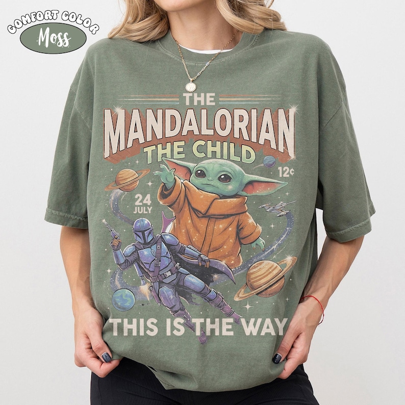 May include: A moss green t-shirt featuring a graphic print of The Mandalorian and The Child in space. The design includes planets, stars, and the text THE MANDALORIAN, THE CHILD, 24 JULY, 12c, and THIS IS THE WAY. A small oval label reads COMFORT COLOR MOSS.