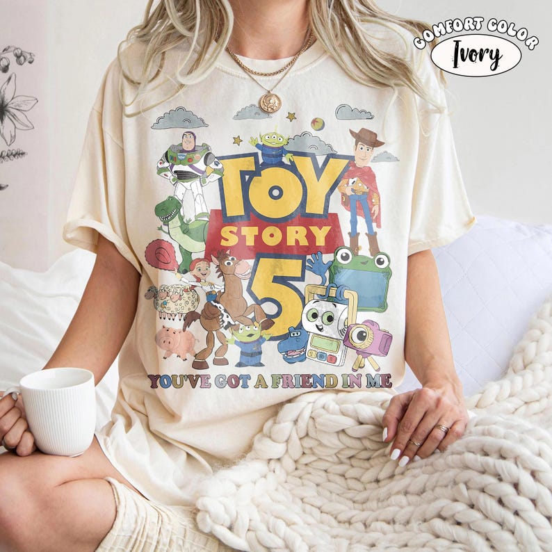 May include: An ivory t-shirt featuring a colorful graphic print of Toy Story characters including Woody, Buzz Lightyear, and Rex. The text reads Toy Story 5 and You've Got A Friend In Me. A small circular label in the corner says Comfort Color Ivory.