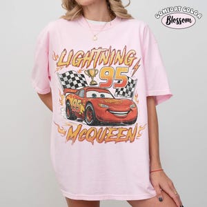 Puede incluir: Una camiseta rosa claro con un gr&aacute;fico de un coche de carreras rojo con el n&uacute;mero 95, un trofeo dorado y banderas a cuadros. El texto dice Lightning, 95 y McQueen, rodeado de llamas naranjas. En la esquina superior derecha dice Comfort Color Blossom.
