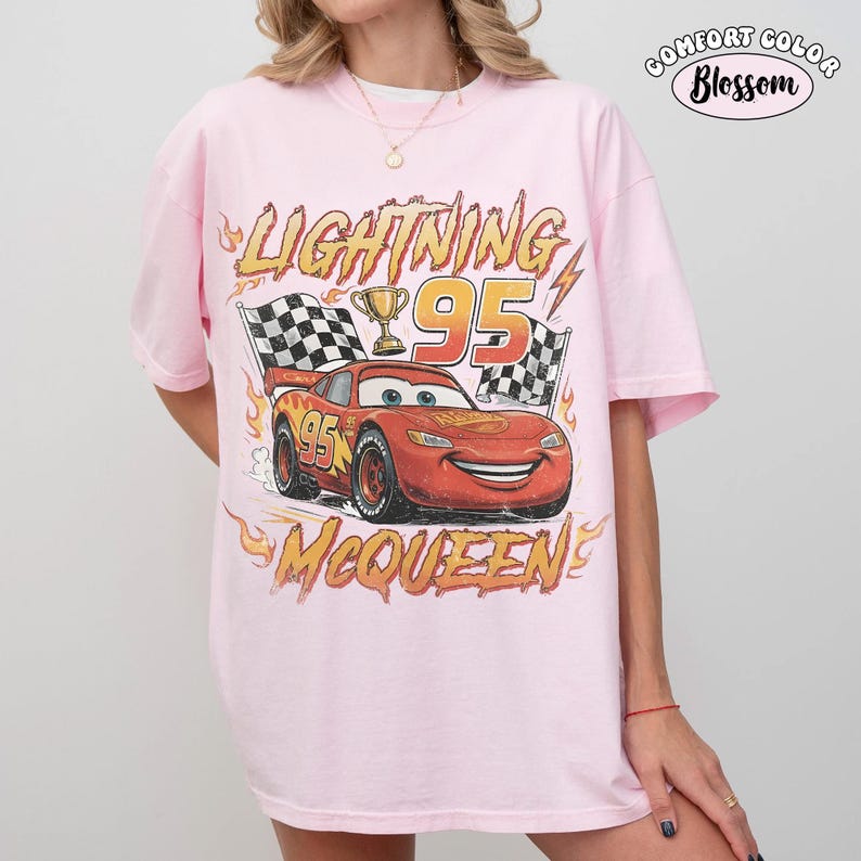 Puede incluir: Una camiseta rosa claro con un gr&aacute;fico de un coche de carreras rojo con el n&uacute;mero 95, un trofeo dorado y banderas a cuadros. El texto dice Lightning, 95 y McQueen, rodeado de llamas naranjas. En la esquina superior derecha dice Comfort Color Blossom.