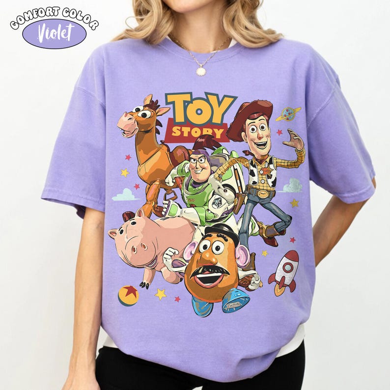 May include: A Comfort Colors violet t-shirt featuring a colorful graphic of characters from the animated film "Toy Story." The design includes Woody, Buzz Lightyear, and other characters. The text "TOY STORY" is displayed prominently.