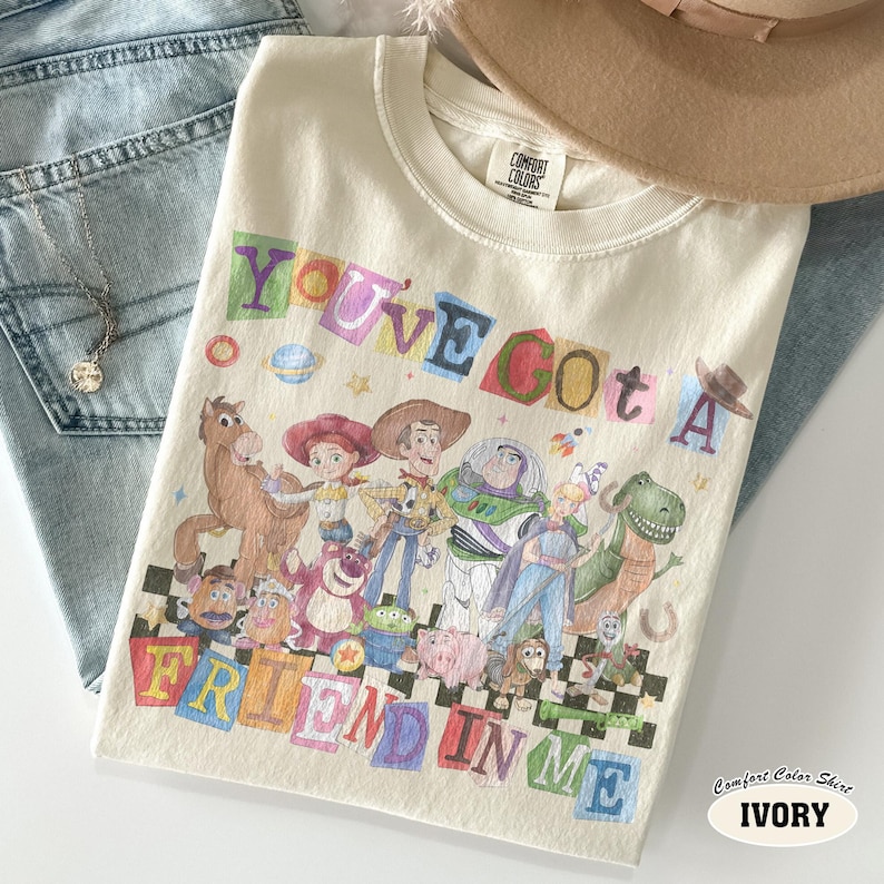May include: Ivory-colored t-shirt featuring a colorful graphic of characters from a popular animated movie, with the text "YOU'VE GOT A FRIEND IN ME." The shirt is paired with blue jeans and a necklace.