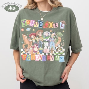 May include: Olive green t-shirt with the text "YOU'VE GOT A FRIEND IN ME" in colorful block letters. The shirt features a graphic of characters from the movie Toy Story, including Woody, Buzz Lightyear, and others. The shirt is labeled "Comfort Color Moss".