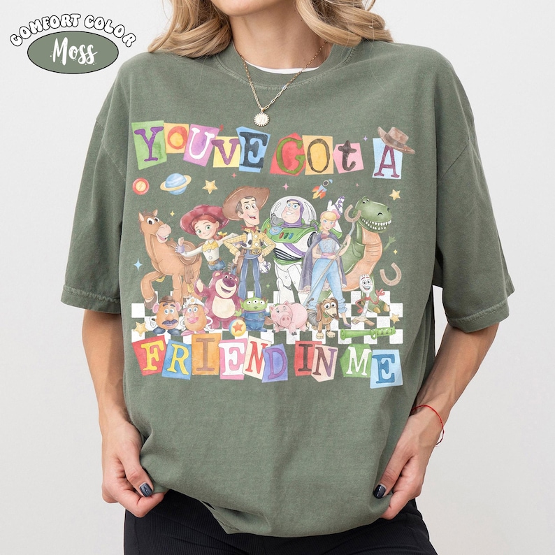 May include: Olive green t-shirt with the text "YOU'VE GOT A FRIEND IN ME" in colorful block letters. The shirt features a graphic of characters from the movie Toy Story, including Woody, Buzz Lightyear, and others. The shirt is labeled "Comfort Color Moss".