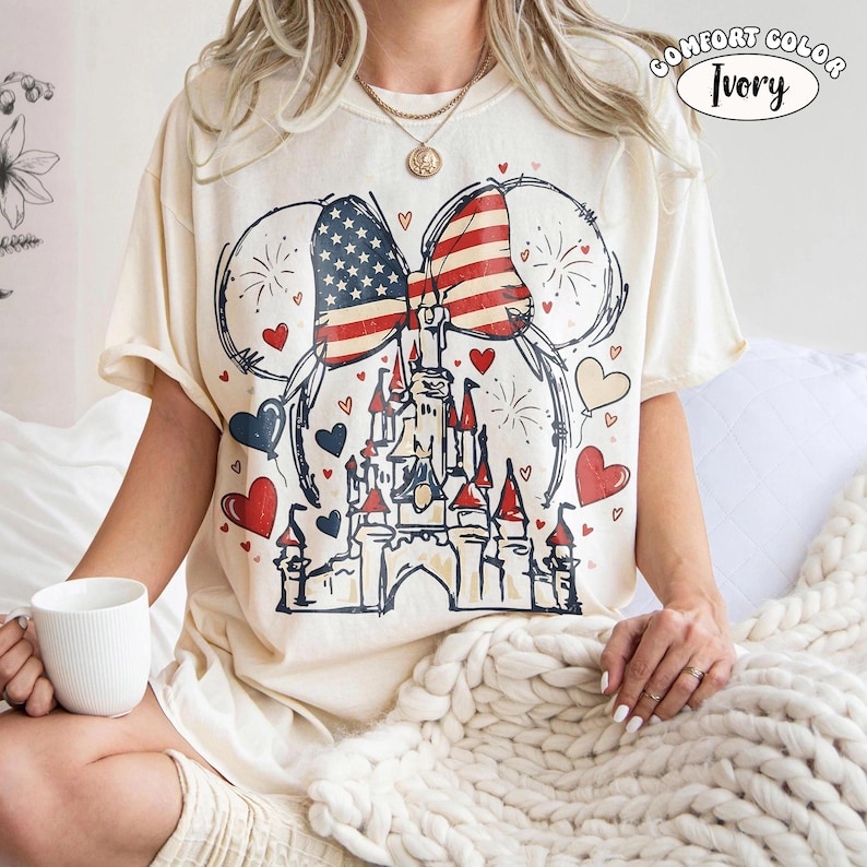 May include: Ivory Comfort Colors t-shirt featuring a patriotic design. The graphic includes a castle, a bow with the American flag, Mickey Mouse ears, hearts, and fireworks. The shirt is short-sleeved and has the words "Comfort Color Ivory" printed on it.