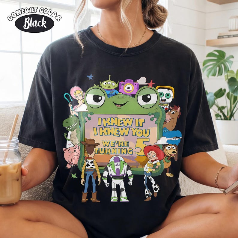 Puede incluir: Una camiseta negra de manga corta con un estampado gr&aacute;fico de varios personajes animados rodeando un marco verde con forma de rana. El texto dentro del marco dice: "I KNEW IT I KNEW YOU WE'RE TURNING 5". Una peque&ntilde;a etiqueta en la esquina dice "COMFORT COLOR Black".
