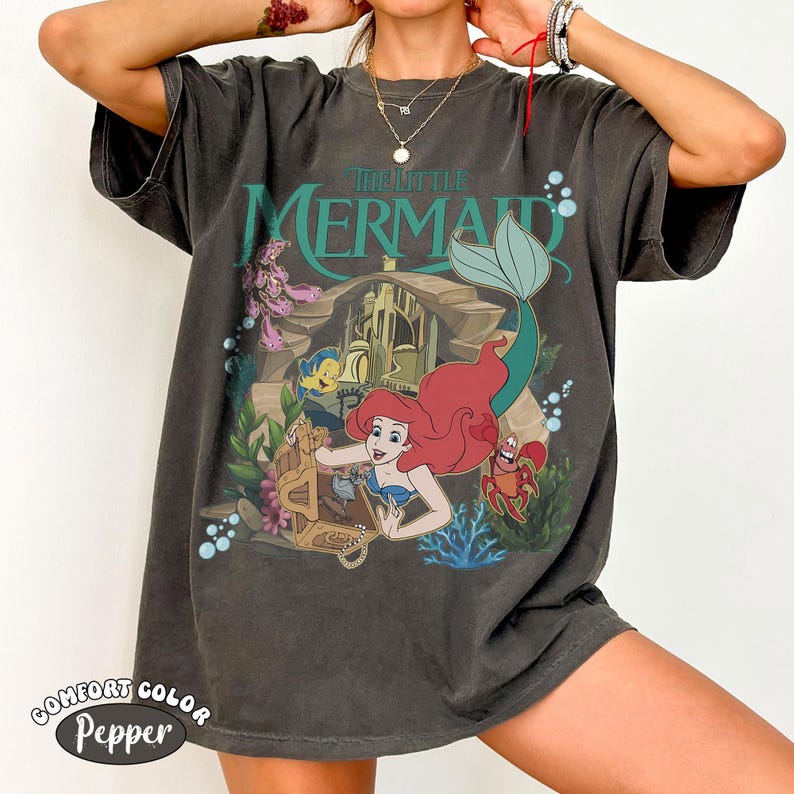 K&ouml;nnte beinhalten: Dunkelgraues T-Shirt mit einer farbenfrohen Grafik von "Arielle, die Meerjungfrau". Das Design zeigt Arielle, Fabius, Sebastian und eine Schatzkiste. Der Text "The Little Mermaid" ist gr&uuml;n. Das Shirt hat den Text "Comfort Color Pepper" unten.