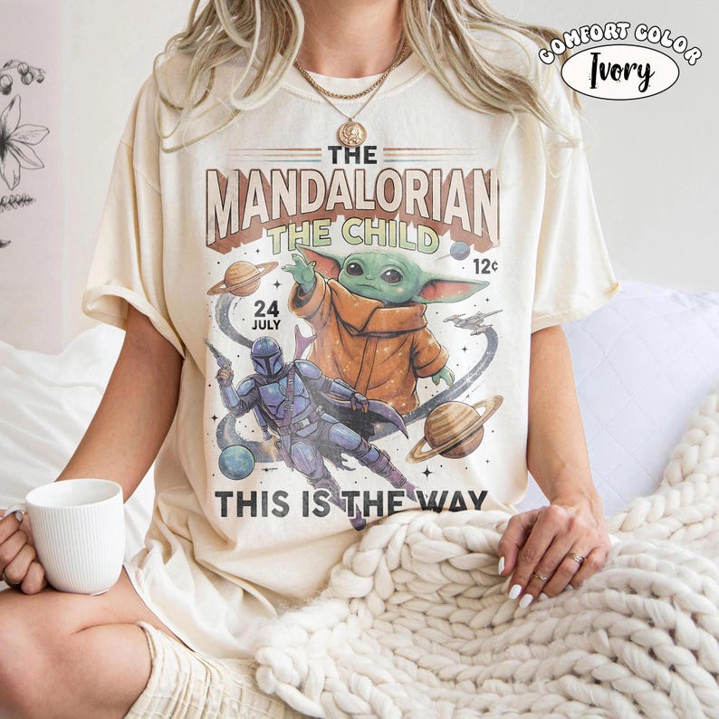 May include: An ivory short-sleeve t-shirt featuring a graphic print of The Mandalorian and The Child in space. The design includes planets, stars, and the text "THE MANDALORIAN THE CHILD", "24 JULY", "12c", and "THIS IS THE WAY" in a vintage style.