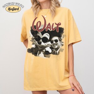 Puede incluir: Camiseta de color mostaza con un gr&aacute;fico de Mickey Mouse con gafas de sol y Walt Disney. La palabra "Walt" est&aacute; impresa encima del gr&aacute;fico en color granate. La camiseta es de la marca Comfort Color.