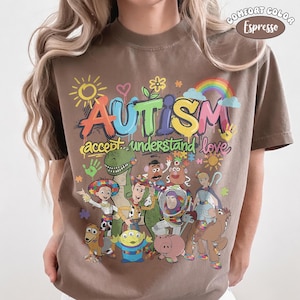 May include: An espresso brown t-shirt featuring a colourful graphic with the text "AUTISM accept understand love" and various cartoon characters including a cowboy, space ranger, cowgirl, dinosaur, and other toys, accented with a sun and rainbow.