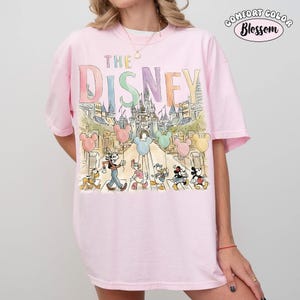 Pu&ograve; includere: T-shirt rosa chiaro con una grafica colorata con la scritta "THE DISNEY" sopra una scena del castello e personaggi Disney. La maglietta ha anche le parole "COMFORT COLOR Blossom" in un cerchio.