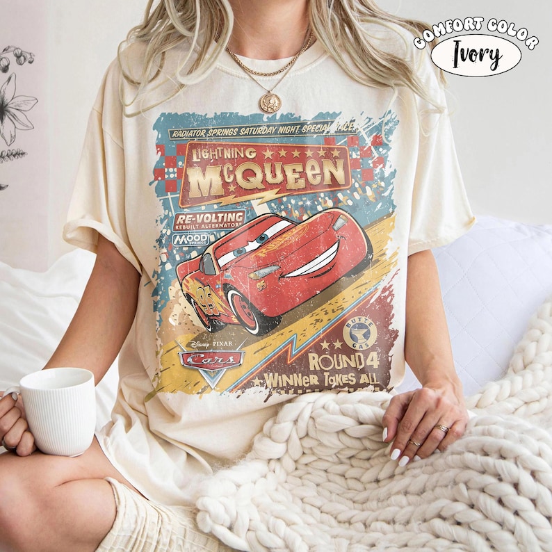May include: Ivory-colored t-shirt featuring a graphic of Lightning McQueen from the Disney Pixar Cars movie. The design includes text that reads "Lightning McQueen" and "Radiator Springs Saturday Night Special". The shirt is a Comfort Color.