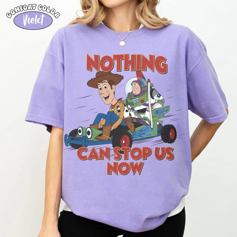 May include: A violet Comfort Colors t-shirt featuring a graphic of Woody and Buzz Lightyear in a green vehicle. The text "NOTHING CAN STOP US NOW" is printed in red. The shirt has short sleeves.