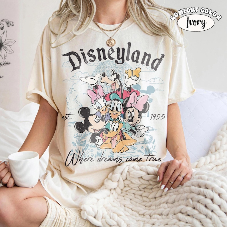 以下が含まれることがあります： アイボリーのTシャツで、ミッキーマウス、ミニーマウス、ドナルドダック、デイジーダック、グーフィー、プルートのグラフィックが描かれています。テキストには「Disneyland」、「est. 1955」、「Where dreams come true」とあります。隅の小さな円形ラベルには「Comfort Color Ivory」と記されています。