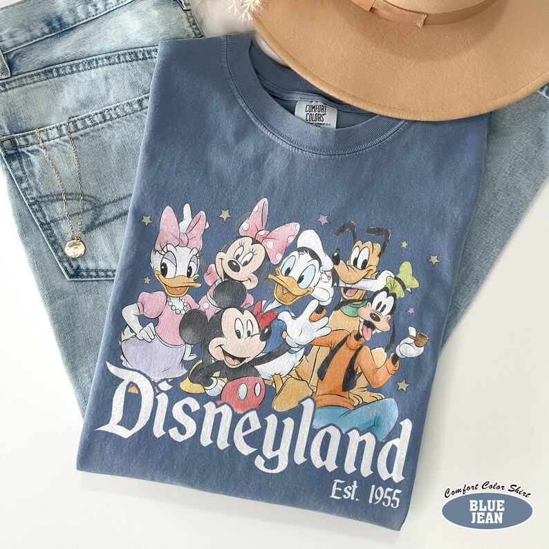 Pu&ograve; includere: Una t-shirt blu a girocollo con una stampa grafica di Topolino, Minnie, Paperino, Paperina, Pluto e Pippo. Il testo Disneyland Est. 1955 &egrave; stampato sotto i personaggi. La maglietta presenta un'etichetta del marchio Comfort Colors.