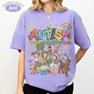 May include: A violet t-shirt featuring a graphic print with the text "AUTISM accept understand love" in colourful letters. The design includes illustrations of various animated toy characters, a sun, a rainbow, and puzzle pieces.