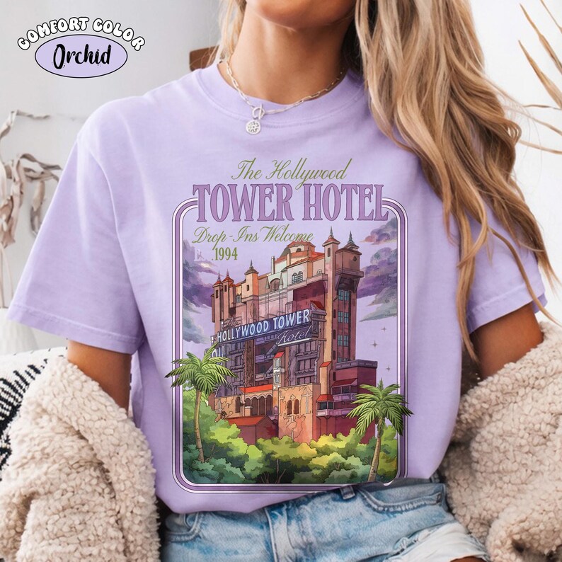 Op de afbeelding: Een lichtpaars T-shirt met een afbeelding van het Hollywood Tower Hotel. Het ontwerp bevat de tekst "The Hollywood Tower Hotel" en "Drop-Ins Welcome 1994". De afbeelding toont een gedetailleerde illustratie van het hotel met palmbomen.