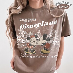 May include: An espresso brown t-shirt featuring a white graphic print of Mickey and Minnie Mouse. The text reads CALIFORNIA Est. 1955 Disneyland The happiest place on earth. A small oval badge in the top right corner says COMFORT COLOR Espresso.