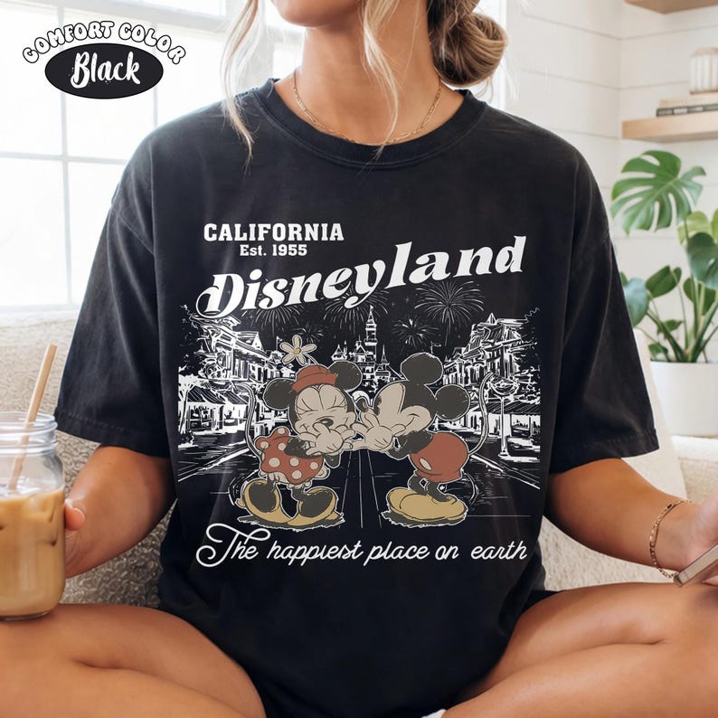 May include: A black short-sleeve t-shirt featuring a white graphic print of Mickey and Minnie Mouse. The text reads California Est. 1955, Disneyland, and The happiest place on earth. The design includes a detailed illustration of the park.
