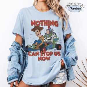 May include: Light blue t-shirt featuring a graphic of Woody and Buzz Lightyear in a vehicle, with the text "NOTHING CAN STOP US NOW" in red. The shirt has rolled-up sleeves and a "Comfort Color Chambray" label.