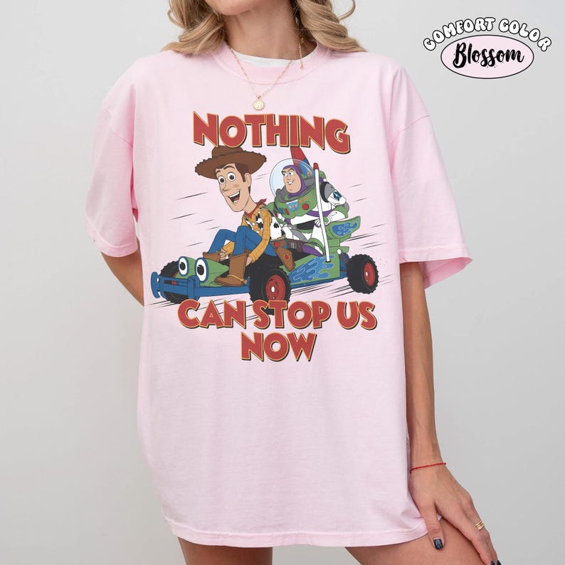 May include: Pink Comfort Colors t-shirt featuring a graphic of Woody and Buzz Lightyear in a race car with the text "NOTHING CAN STOP US NOW." The shirt has short sleeves and a crew neck.
