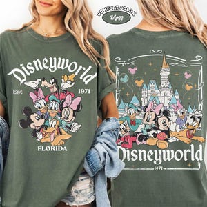 K&ouml;nnte beinhalten: Ein moosgr&uuml;nes T-Shirt mit einer Vorderseite, die den Schriftzug Disneyworld, Est 1971, Florida und Zeichentrickfiguren zeigt. Die R&uuml;ckseite zeigt ein Schloss, den Schriftzug Disneyworld 1971 und Figuren mit Luftballons in einem Rahmen.