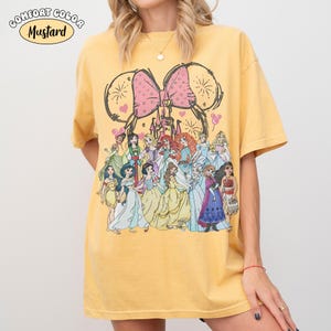Op de afbeelding: Mosterdkleurig T-shirt met een afbeelding van Disney-prinsessen. Het ontwerp bevat een grote roze strik, kasteel en muisoren. De tekst "Comfort Color" en "Mustard" staat linksboven. Het shirt is een comfortabele, casual stijl.