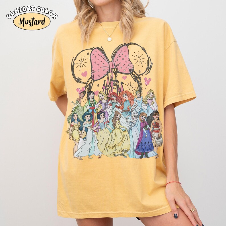 Op de afbeelding: Mosterdkleurig T-shirt met een afbeelding van Disney-prinsessen. Het ontwerp bevat een grote roze strik, kasteel en muisoren. De tekst "Comfort Color" en "Mustard" staat linksboven. Het shirt is een comfortabele, casual stijl.