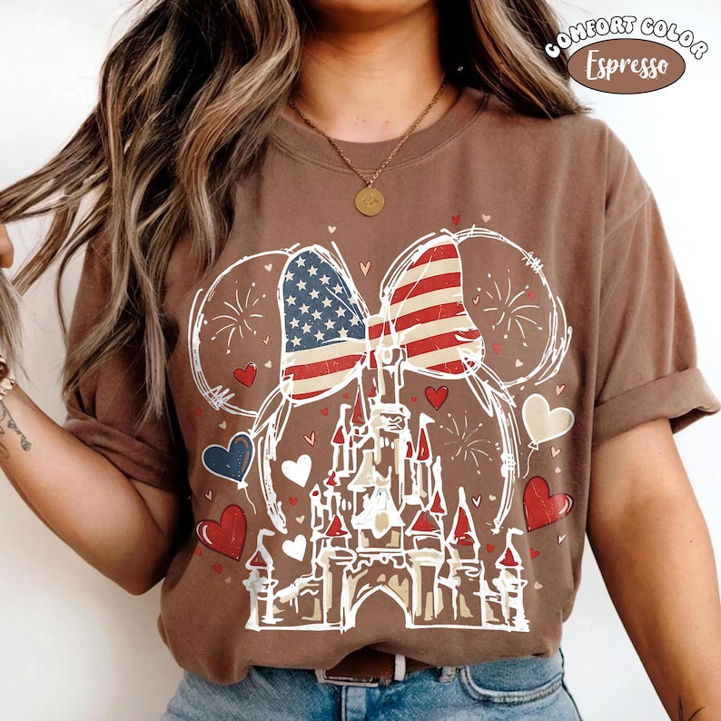 May include: Brown t-shirt with a graphic featuring a castle, hearts, and a bow designed with the American flag. The shirt has the words "Comfort Color Espresso" printed on the top right corner.