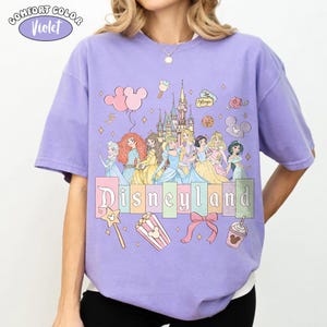 Puede incluir: Camiseta violeta Comfort Colors con un gr&aacute;fico de Disneyland. El dise&ntilde;o incluye princesas de Disney, un castillo y la palabra "Disneyland" en un estilo de letras de bloque colorido. Gr&aacute;ficos adicionales incluyen globos, palomitas de ma&iacute;z y un lazo.