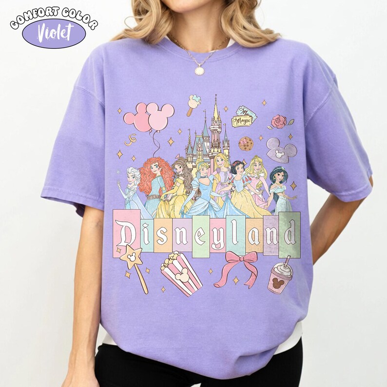 Puede incluir: Camiseta violeta Comfort Colors con un gr&aacute;fico de Disneyland. El dise&ntilde;o incluye princesas de Disney, un castillo y la palabra "Disneyland" en un estilo de letras de bloque colorido. Gr&aacute;ficos adicionales incluyen globos, palomitas de ma&iacute;z y un lazo.
