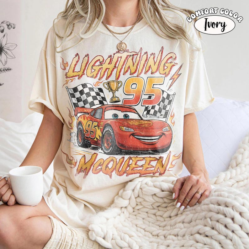 Puede incluir: Una camiseta de manga corta color marfil con un gr&aacute;fico de un coche de carreras rojo con el n&uacute;mero 95, un trofeo y banderas a cuadros. El texto dice LIGHTNING 95 MCQUEEN. Una etiqueta circular en la esquina superior derecha muestra COMFORT COLOR IVORY.
