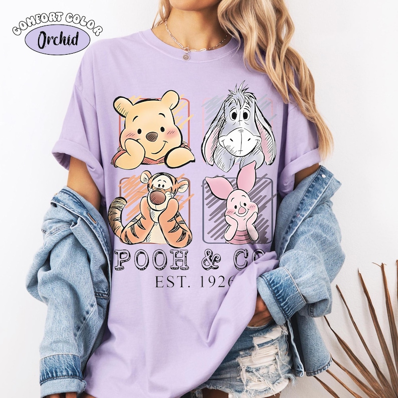 May include: A lilac Comfort Colors t-shirt featuring four cartoon characters: Winnie the Pooh, Eeyore, Tigger, and Piglet, each in a square frame. The text "POOH & CO. EST. 1926" is printed below the characters. The shirt has short sleeves.
