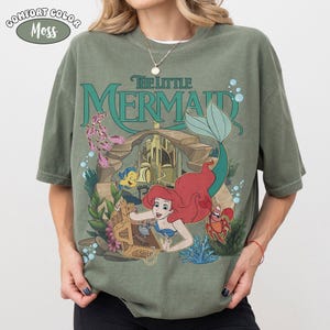K&ouml;nnte beinhalten: Moosgr&uuml;nes T-Shirt mit einem Aufdruck von Arielle aus Arielle, die Meerjungfrau. Das Design umfasst den Filmtitel, Arielle, Fabius, Sebastian und Unterwasserelemente. Das Shirt ist von der Marke Comfort Colors.