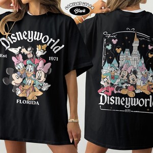 K&ouml;nnte beinhalten: Ein schwarzes T-Shirt mit einer Vorderseite, die Mickey Mouse, Minnie Mouse, Donald Duck, Daisy Duck, Goofy und Pluto mit dem Text Disneyworld Est 1971 Florida zeigt. Die R&uuml;ckseite zeigt dieselben Figuren vor einem Schloss mit Luftballons.