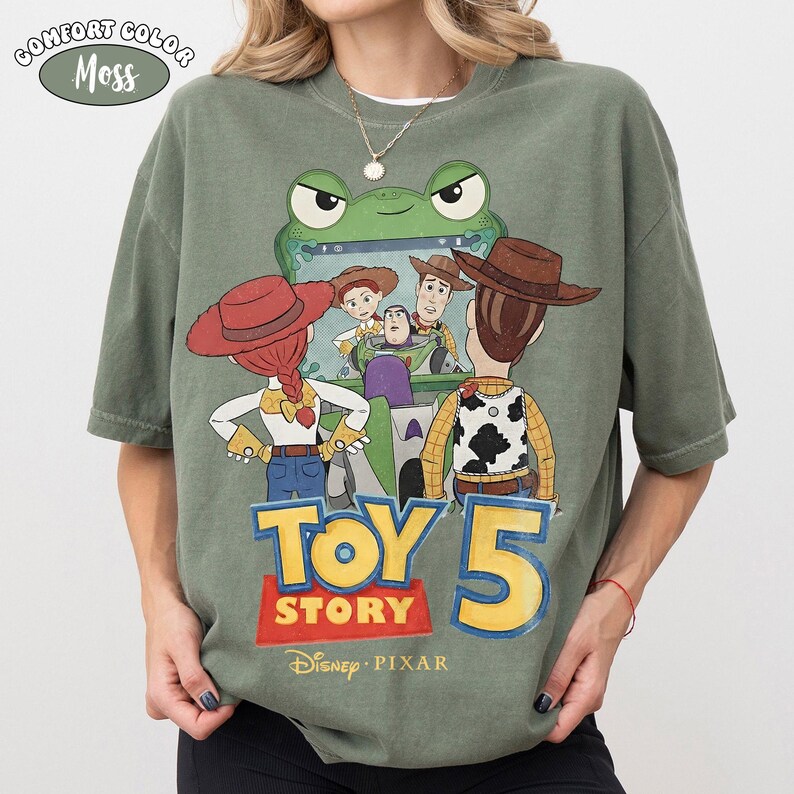 Pu&ograve; includere: T-shirt Comfort Colors verde oliva con grafica di Toy Story 5. Il design presenta Woody, Buzz Lightyear e Jessie, con "TOY STORY 5" in giallo e rosso. &Egrave; visibile anche il logo Disney Pixar.