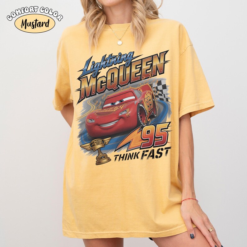 May include: Mustard-colored t-shirt featuring Lightning McQueen from the Cars movie. The graphic includes the character, the number 95, and the words "THINK FAST". The shirt also has the words "COMFORT COLOR" and "Mustard" printed on it.