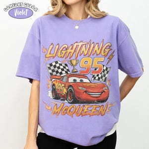 Puede incluir: Una camiseta violeta de manga corta con un gr&aacute;fico del coche de carreras rojo Lightning McQueen, el n&uacute;mero 95, un trofeo dorado y banderas a cuadros. El texto dice LIGHTNING 95 MCQUEEN con llamas naranjas. Se ve una etiqueta Comfort Color Violet.