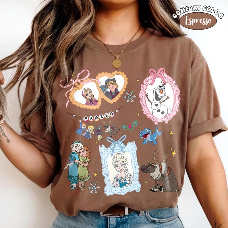 May include: Espresso-colored t-shirt featuring various characters and scenes from the animated film "Frozen." The design includes framed portraits of characters, snowflakes, and the word "Frozen" in a decorative font. The shirt is a Comfort Color brand.