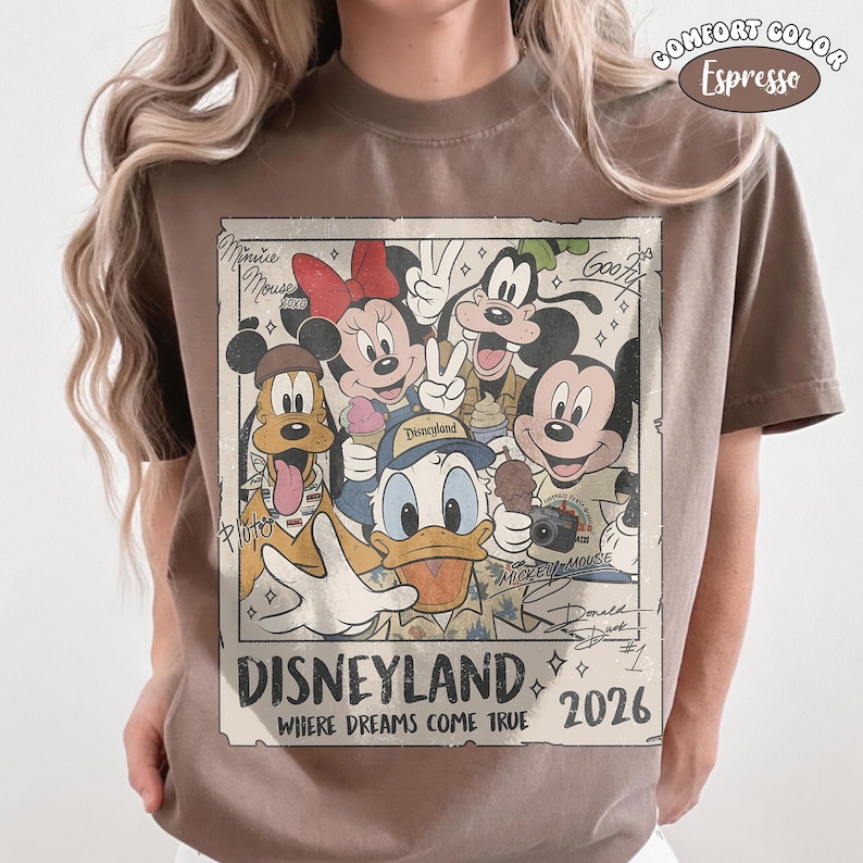 Puede incluir: Una camiseta color marr&oacute;n espresso con un gr&aacute;fico rectangular de personajes de Disney que incluye a Mickey Mouse, Minnie Mouse, el Pato Donald, Goofy y Pluto. El texto dice Disneyland, Where Dreams Come True y 2026, con firmas de personajes.