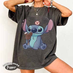 May include: A charcoal gray short-sleeve t-shirt featuring a graphic of the character Stitch wearing a black hat with mouse ears and holding a brown chocolate treat shaped like a mouse head. The shirt has a faded, vintage-style wash finish.