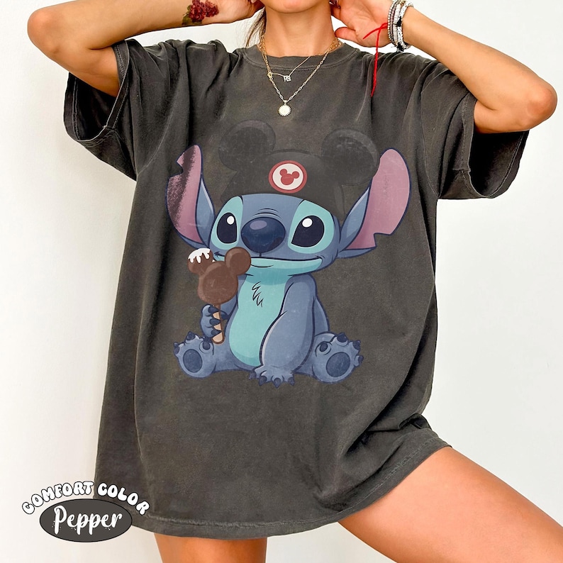 May include: A charcoal gray short-sleeve t-shirt featuring a graphic of the character Stitch wearing a black hat with mouse ears and holding a brown chocolate treat shaped like a mouse head. The shirt has a faded, vintage-style wash finish.