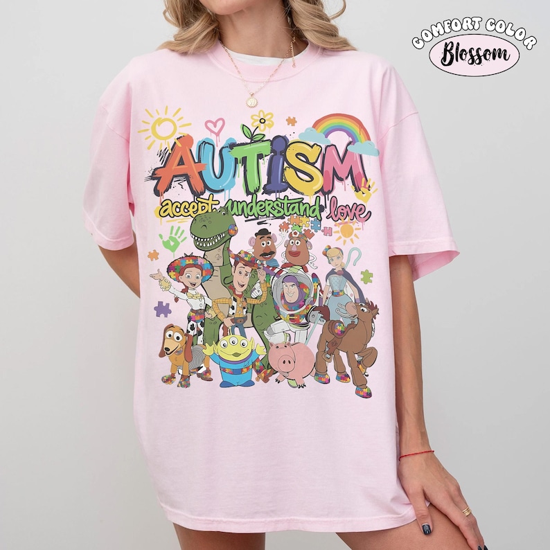 May include: A light pink t-shirt featuring a colourful graphic print of various animated characters. The text reads "AUTISM" in large letters above "accept understand love" in cursive. Additional graphics include a sun, rainbow, and puzzle pieces.