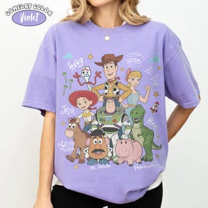 Może przedstawiać: Jasnofioletowy t-shirt z kr&oacute;tkim rękawem z nadrukiem graficznym przedstawiającym postacie z Toy Story, w tym Chudego, Buzza Astrala, Jessie, Rexa, Hamm, Pana Bulwę, Celka i Sztućka, z ich imionami zapisanymi kursywą.