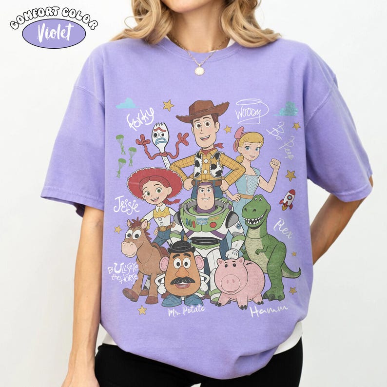 Może przedstawiać: Jasnofioletowy t-shirt z kr&oacute;tkim rękawem z nadrukiem graficznym przedstawiającym postacie z Toy Story, w tym Chudego, Buzza Astrala, Jessie, Rexa, Hamm, Pana Bulwę, Celka i Sztućka, z ich imionami zapisanymi kursywą.