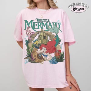 K&ouml;nnte beinhalten: Hellrosa T-Shirt mit einem Motiv aus "Arielle, die Meerjungfrau". Das Design zeigt Arielle, Fabius, Sebastian und eine Schatztruhe. Die Worte "The Little Mermaid" sind gr&uuml;n. Das Shirt hat ein Comfort Colors Blossom-Label.