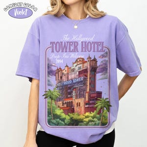 Op de afbeelding: Een violet Comfort Colors T-shirt met een afbeelding van het Hollywood Tower Hotel. Het ontwerp bevat de tekst "The Hollywood Tower Hotel, Drop-Ins Welcome, 1994" en een gestileerde afbeelding van het hotel.